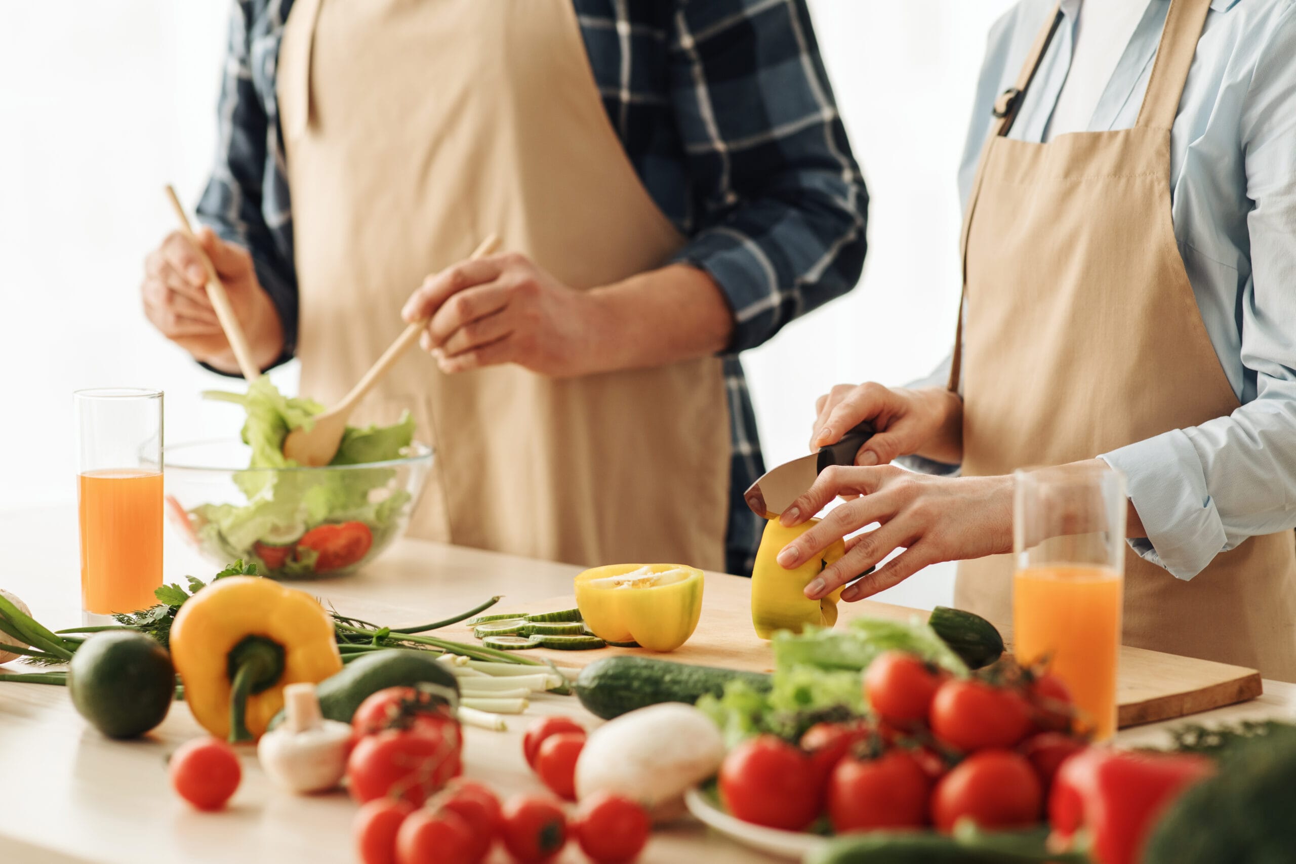 Vegetables for proper nutrition, diet for healthcare. Mature husband and wife in aprons prepare salad in kitchen interior with vegetables and glasses of vitamin juice on table, cropped, close up